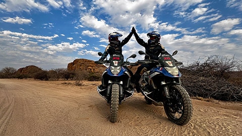 Dos conductoras del equipo femenino francés sentadas una junto a la otra en sus motos de competición R 1300 GS se chocan los cinco.