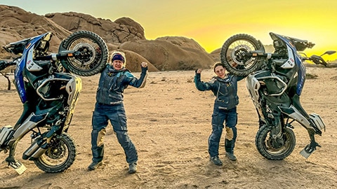Las conductoras del equipo alemán al atardecer junto a sus motos, que solo tocan el suelo con las ruedas traseras. Una de sus manos se apoya en la rueda delantera. Levantan la otra mano cerrada hacia arriba.